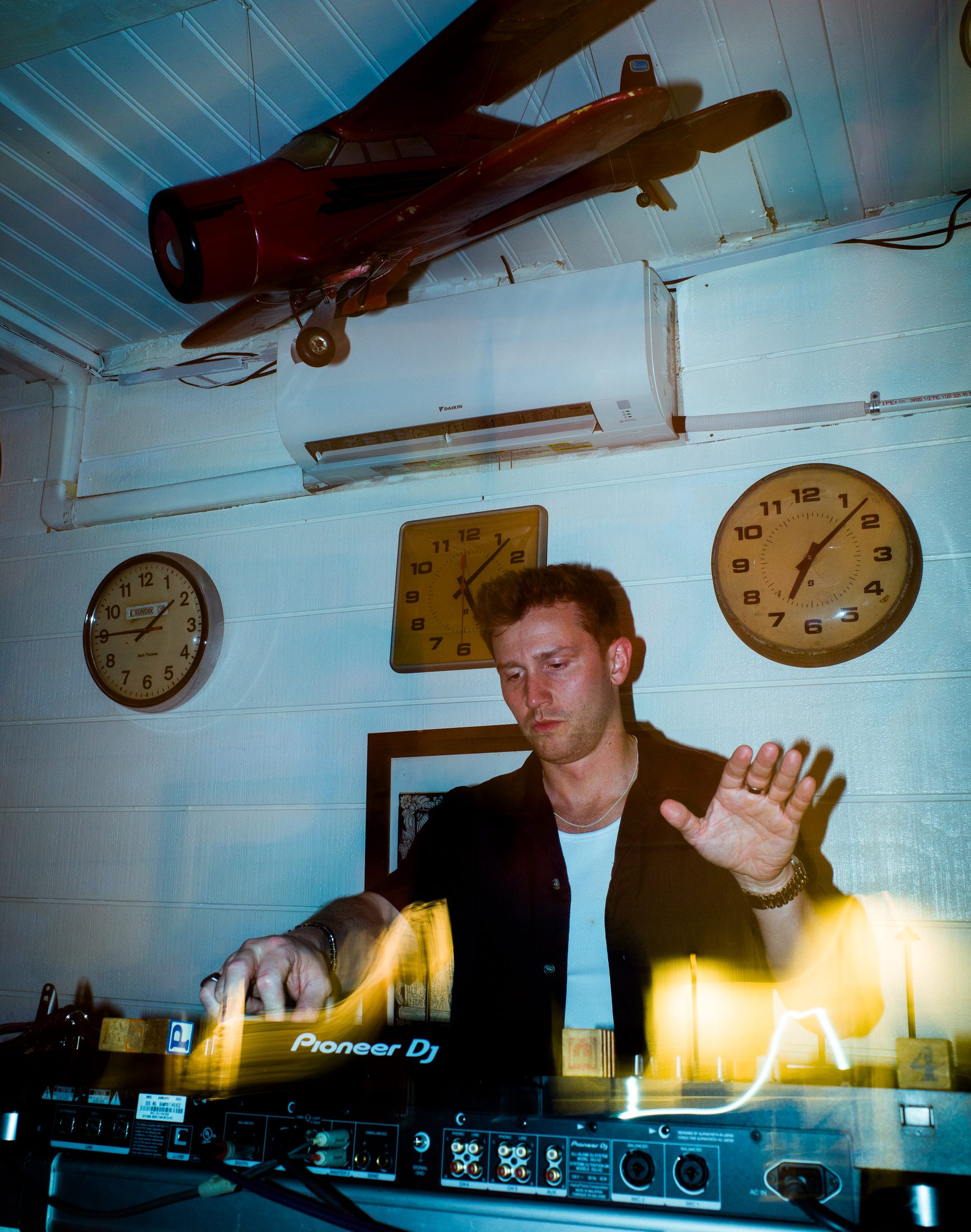 Double-exposure DJ set behind Pioneer decks, vintage wall clocks and a model plane above